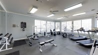 This fitness center offers a welcoming and functional environment designed to support a well-rounded exercise routine. The space is finished with soft gray walls and crown molding, lending a clean, polished aesthetic that sets it apart from a typical gym setting. Durable rubber tile flooring covers the entire area, providing cushioning and traction suitable for a variety of workouts.

Natural light is a defining feature of this fitness center, with multiple large windows fitted with horizontal blinds lining the walls. These windows frame views of surrounding trees, creating a bright and refreshing atmosphere throughout the day. Two ceiling fans are mounted overhead, promoting airflow and comfort during intense sessions.

The equipment selection is comprehensive, encompassing both cardio and strength training options. Cardio machines include elliptical trainers and a stationary bike, while strength equipment features a cable/weight machine and a dedicated free weight area with an adjustable bench and dumbbells. The layout is open and well-organized, allowing users to move freely between stations.

Overall, the fitness center delivers a bright, comfortable, and fully equipped space that supports residents' health and wellness goals conveniently on-site.