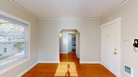 Located at 2256 NW Overton St in Portland, Oregon, this single-floor unit offers a thoughtfully arranged layout comprising a living room, kitchen, hallway, bathroom, laundry room, and closet. The living room is a bright, welcoming space highlighted by warm hardwood flooring, neutral painted walls, white-trimmed double-hung windows that frame views of the surrounding greenery, and a classic cast-iron radiator providing radiant heat. A charming arched doorway connects the living area to the hallway and kitchen, adding a timeless architectural detail to the interior. The kitchen features white cabinetry, a stainless steel sink, rich laminate countertops with a stone-like finish, and tile flooring, with a window that brings in natural light. The laundry room is a practical space equipped with white upper cabinets for storage, a tall closet-style panel, and tile flooring. The bathroom includes a window with frosted glass for privacy, tilework at the base, and clean white fixtures. Throughout, the unit maintains a cohesive, understated aesthetic with neutral tones, white trim, and natural light filtering through multiple windows—creating a comfortable and functional living environment in the heart of Portland's Northwest neighborhood.