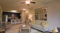 This single-floor residence offers a thoughtfully designed open-concept layout that seamlessly connects the living and kitchen areas, creating a cohesive and functional living environment. The living room serves as the centerpiece of the space, featuring a ceiling fan with light fixture overhead and warm ambient lighting that gives the room an inviting, comfortable feel. A neutral-toned sofa accented with yellow throw pillows anchors the seating area, complemented by a rustic round coffee table. A set of decorative framed mirrors mounted on the wall adds depth and visual interest to the room. The kitchen is outfitted with dark cabinetry, stainless steel appliances including a microwave and oven range, and a breakfast bar with bar-height seating for multiple guests — making it both practical and social. Pendant lighting above the bar adds a refined touch. The interior palette features warm beige and neutral tones throughout, contributing to a cohesive and welcoming ambiance. Light-toned flooring flows through the open space, enhancing the sense of continuity between the living and kitchen areas. The overall design balances functionality with tasteful décor, making this a comfortable and well-presented single-level home.