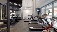 This fitness center is thoughtfully designed to support a comprehensive workout experience within a modern, single-level layout. The space commands attention with soaring double-height ceilings and expansive floor-to-ceiling windows fitted with frosted glass panels, allowing abundant natural light to fill the room while maintaining privacy. A striking spiral staircase adds an architectural focal point and connects to an upper level overlooking the gym floor.

The cardio zone is well-appointed with multiple treadmills, elliptical trainers, stationary bikes — including both upright and recumbent models — and a rowing machine, each equipped with individual monitors for entertainment during workouts. Wall-mounted flat-screen televisions are positioned throughout the space, ensuring visibility from all exercise stations.

Large mirror panels line portions of the walls, visually expanding the space and supporting proper form during exercise. The flooring consists of durable, dark-toned carpet tiles suited for fitness use. A hydration station with dual water fountains — including an accessible-height unit — is conveniently located near the entrance. Recessed lighting complements the natural light, maintaining a well-lit atmosphere throughout. The overall design is clean, functional, and modern, offering an inviting environment for daily fitness routines.