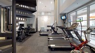 This fitness center is thoughtfully designed to support a comprehensive workout experience within a modern, single-level layout. The space commands attention with soaring double-height ceilings and expansive floor-to-ceiling windows fitted with frosted glass panels, allowing abundant natural light to fill the room while maintaining privacy. A striking spiral staircase adds an architectural focal point and connects to an upper level overlooking the gym floor.

The cardio zone is well-appointed with multiple treadmills, elliptical trainers, stationary bikes — including both upright and recumbent models — and a rowing machine, each equipped with individual monitors for entertainment during workouts. Wall-mounted flat-screen televisions are positioned throughout the space, ensuring visibility from all exercise stations.

Large mirror panels line portions of the walls, visually expanding the space and supporting proper form during exercise. The flooring consists of durable, dark-toned carpet tiles suited for fitness use. A hydration station with dual water fountains — including an accessible-height unit — is conveniently located near the entrance. Recessed lighting complements the natural light, maintaining a well-lit atmosphere throughout. The overall design is clean, functional, and modern, offering an inviting environment for daily fitness routines.