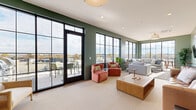 Located at 2100 Dabney Rd in Richmond, Virginia, this single-floor living room lounge offers a thoughtfully designed communal space defined by its modern aesthetic and abundant natural light. The room is enveloped in sage and deep forest green walls, creating a calm, inviting atmosphere that complements the neutral carpeted flooring throughout.

Floor-to-ceiling black-framed windows and a full-height glass door dominate one wall, flooding the interior with daylight and providing sweeping outdoor views of the surrounding Richmond skyline and neighborhood. The glass door opens directly onto a furnished terrace, extending the living space outdoors with additional seating for relaxing or entertaining.

Inside, the lounge is thoughtfully arranged with multiple seating areas. A tufted leather sofa and a sectional gray couch anchor the central gathering zone around a solid wood coffee table. Warm terracotta-toned accent chairs provide colorful contrast, while layered area rugs add texture and define each seating cluster. A wall-mounted flat-screen television, a sleek media console, and framed botanical artwork complete the space with polished, residential-style details.

Recessed ceiling lighting and a pendant fixture ensure the room remains well-lit at all hours. Lush potted plants are positioned throughout, enhancing the organic, welcoming character of the lounge. The overall design balances comfort and contemporary style, making it an ideal space for gathering and relaxation.