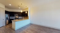 This single-floor residence is designed with a contemporary aesthetic and an open-concept layout that promotes a seamless flow between the kitchen and living areas. The kitchen serves as a central highlight, featuring dark espresso cabinetry that extends to the ceiling for generous storage, granite countertops, and a taupe subway tile backsplash. Stainless steel appliances — including a refrigerator, range, and microwave — complement the modern design. A peninsula with a granite surface and an undermount sink adds both functionality and a natural dividing point between the kitchen and the expansive living area. Pendant lighting above the peninsula adds a warm, focused ambiance to the space.

The living area is generously proportioned, finished with wood-style flooring and crisp white walls that enhance brightness. A ceiling fan and a glass-panel door leading to a private balcony introduce natural light and outdoor views. Built-in shelving is also present, offering additional display and storage options.

The bathroom mirrors the kitchen's design language, with a dark vanity, granite countertop, framed mirror, and tile flooring. A spacious walk-in closet features multiple hanging rods and shelving, providing substantial storage capacity throughout the unit.