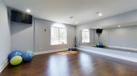 Located at 700 Citation Lane in Cherry Hill, NJ, this single-floor fitness studio is thoughtfully designed to support a variety of workout routines. The space features rich, dark hardwood flooring that provides both durability and an attractive aesthetic underfoot. Large arched and rectangular windows allow generous natural light to flood the room, creating a bright and energizing environment for exercise. A full-length wall mirror spans a significant portion of one wall, offering exercisers the ability to monitor their form during workouts while also enhancing the sense of spaciousness. The walls are finished in a clean, neutral gray tone that lends a calm and focused atmosphere. Recessed lighting complements the natural light for consistent illumination throughout. Fitness accessories are neatly organized within the space, including a medicine ball rack stocked with weighted balls of varying sizes and stability balls positioned along the walls. A mounted flat-screen television adds a multimedia element for guided workouts or entertainment. The open floor plan allows for flexible use, accommodating yoga, stretching, aerobics, and strength training activities. The overall design balances functionality with a clean, modern aesthetic, making it a well-equipped and inviting space for fitness enthusiasts.