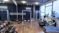 This single-floor fitness studio is thoughtfully designed to support a comprehensive workout experience. The space features a bold, modern aesthetic defined by deep navy acoustic panel walls accented with gray tones, creating a focused and motivating atmosphere. Durable hardwood-style flooring runs throughout, providing both functionality and visual warmth.

The studio is outfitted with a range of professional-grade equipment, including a multi-functional power rack and cable machine for strength training, a flat adjustable weight bench, a set of dumbbells, and foam rollers for recovery work. On the cardio side, an elliptical trainer is prominently positioned near a large window, which allows natural light to fill the space and provides an open, energizing feel.

Overhead, a combination of flush and track lighting ensures the entire floor is well-illuminated for safe and effective training. A full-length mirror along one wall enhances the sense of space while allowing users to monitor their form. An inspirational wall graphic reinforces the motivational tone of the environment. This studio is well-suited for both individual training sessions and small-group workouts, offering a versatile and professionally appointed fitness environment.