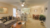 This single-floor residence offers an open-concept layout that seamlessly connects the living room, dining area, and kitchen into one cohesive and functional living space. The living room is anchored by a plush sofa and a classic wooden coffee table, with a ceiling fan overhead providing both comfort and a traditional aesthetic. Natural light filters in through a window dressed with blinds, brightening the warm, neutral tones of the carpet that flows throughout the space. A raised breakfast bar with elegant wrought-iron barstools serves as a natural dividing element between the living area and the kitchen, offering casual seating and an open sightline throughout the floor plan. The kitchen is well-appointed with white cabinetry, a full refrigerator, and ample counter space, designed for both everyday cooking and entertaining. Adjacent to the kitchen, the dining area accommodates a full-sized wooden table with seating for six, lit warmly from above by a decorative chandelier. The overall ambiance is cozy and welcoming, characterized by warm earth tones, soft carpeting, and a cohesive design that makes the space feel both functional and homely.