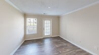 Located at 601 Downsby Ln in Woodstock, Georgia, this two-bedroom, one-bathroom home offers a practical and comfortable single-story layout. The combined kitchen and living area serves as the heart of the home, featuring durable hardwood-style flooring, crown molding, and a textured ceiling. The kitchen is equipped with white cabinetry, granite-look laminate countertops, a double-basin sink, white appliances including a range and refrigerator, and a breakfast bar that connects seamlessly to the living space. A glass-panel door in the living room leads to an outdoor area, while a window allows natural light to fill the room. Both bedrooms are carpeted and feature ceiling fans, large windows that bring in natural light, and panel doors. The primary bedroom is generously sized and includes a walk-in closet outfitted with wire shelving and hanging rods for ample storage. The full bathroom features a wide vanity with a large mirror and Hollywood-style lighting. A dedicated laundry area with washer/dryer hookups and a wire shelving unit adds everyday convenience. Neutral warm-beige tones and white trim carry throughout, creating a cohesive and inviting atmosphere across the entire home.