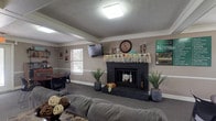 Located at 4121 Kimberly Parkway N in Columbus, Ohio, this single-floor space offers a warm and inviting atmosphere designed for comfort and community gathering. The main lounge area serves as the focal point of the space, centered around a striking dark-painted fireplace that anchors the room with a classic, cozy charm. The mantle is decorated with a wall clock, candle holders, and seasonal greenery, adding a lived-in, welcoming character to the space.

The room features neutral-toned walls accented by clean white crown molding and a wainscoting-style chair rail, lending a polished, traditional aesthetic. Carpet flooring in a muted tone runs throughout, contributing to the room's comfortable feel. Overhead, a textured ceiling is lit by a flush-mount ceiling light, ensuring the space is well-illuminated.

Seating includes a plush gray sofa and a separate dining or meeting area with a dark wood table and chairs positioned near a window, allowing for natural light to filter in. Potted greenery placed on either side of the fireplace adds a fresh, decorative touch. A wall-mounted flat-screen television provides a functional amenity for residents or guests. Overall, the space communicates a cozy, community-oriented environment with thoughtful finishing details throughout.