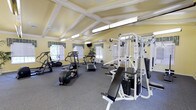 This fitness center is designed to support a well-rounded exercise routine in a bright, comfortable setting. The space is defined by its vaulted ceiling with distinctive exposed white beams, which lend an open, airy quality to the room. Soft yellow walls and multiple windows dressed with patterned valances allow natural light to filter in, creating an inviting atmosphere. Ceiling fans are mounted between the beams, ensuring adequate airflow throughout the space. The flooring consists of durable grey carpet, well-suited for a high-traffic fitness environment. The facility is comprehensively equipped with a variety of cardio machines, including treadmills, an elliptical trainer, and a stair climber, all arranged to allow comfortable use. A multi-station cable weight machine serves as the centerpiece for strength training, accompanied by a dedicated adjustable bench. A wall-mounted television is positioned for visibility from multiple workout stations, adding to the overall convenience. Lush potted plants are placed throughout the room, softening the functional aesthetic and contributing to a pleasant, energizing environment. The thoughtful layout and abundance of equipment make this fitness center a practical and well-appointed facility for residents or guests.