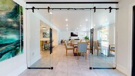 This single-floor space is designed with an open, connected layout that seamlessly integrates multiple functional zones. Entry is made through a standout architectural feature: full-height glass panel barn doors mounted on industrial black hardware, creating a transparent yet defined threshold that immediately conveys the space's modern, welcoming character.

Inside, large-format light stone tile flooring extends throughout, unifying the dining, lounge, and kitchen areas. The dining zone accommodates multiple tables surrounded by wood-framed upholstered chairs, positioned to take advantage of generous natural light from several windows overlooking lush greenery outdoors. A wall-mounted flat-screen television anchors the lounge area, which features a bold textured black fireplace surround, a cushioned sectional sofa, a solid wood coffee table, and a striped area rug layered over the tile.

The kitchen is outfitted with warm natural wood cabinetry, white quartz countertops, stainless steel appliances including a double wall oven, and an eye-catching handmade ceramic tile backsplash in soft blue-white tones. Recessed lighting throughout ensures consistent, even illumination, complementing the crisp white walls and creating a bright, clean ambiance across the entire space.