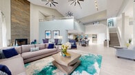 Located at 7610 W Waters Ave in Tampa, FL, this well-appointed two-story clubhouse offers generous square footage across both levels, designed to serve as a vibrant communal gathering space. The ground floor welcomes visitors with an expansive open layout featuring large-format light tile flooring, soaring double-height ceilings, and a grand staircase that anchors the central common area. A fully equipped kitchen showcases white shaker cabinetry, dark granite countertops, stainless steel appliances, and a breakfast bar with seating — all bathed in natural light from large windows overlooking the pool area. The first floor also includes distinct lounge seating areas adorned with contemporary artwork in cool blue and teal tones, a decorative wood-paneled accent wall with a mounted flat-screen TV, and dedicated workspace with computer stations. Upstairs, the second floor features vaulted ceilings with skylights that flood the space with natural light, a full billiards table, a shuffleboard table, and a high-top seating nook beside oversized windows with outdoor views. Sliding glass doors open to a furnished balcony, extending the living space outdoors. Throughout, a cohesive coastal-inspired palette of blues, grays, and neutrals creates a bright, welcoming ambiance.