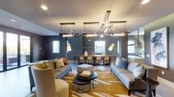 This clubroom offers a sophisticated and comfortable communal gathering space designed with a modern aesthetic. The open layout seamlessly integrates a lounge area and a dining area, making it well-suited for both casual socializing and organized events. Deep charcoal-gray walls create a bold, contemporary backdrop that contrasts elegantly with warm-toned furnishings throughout the space.

The lounge area is anchored by a pair of generously sized gray sofas and a plush accent chair in a warm neutral tone, all arranged around a round nesting coffee table set atop a striking gold and ivory patterned area rug. Accent pillows in rust orange and muted tones add warmth and visual interest to the seating arrangement, while a square upholstered ottoman serves as a functional centerpiece.

Overhead, a series of linear wood-and-metal pendant fixtures and an angular chandelier provide layered ambient and task lighting, complemented by recessed ceiling lights and tall adjustable floor lamps. Natural light filters in through multiple windows, enhancing the welcoming atmosphere.

The dining area features a long communal table with ample seating, positioned beneath a window that offers a view of greenery outside, reinforcing the space's bright and inviting character.