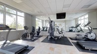 This fitness center offers a well-organized and functional workout environment designed to support a range of exercise routines. The space features light-toned tile flooring with protective rubber mats positioned beneath the equipment to ensure safety and durability. A variety of machines are available, including stationary bikes, a multi-station cable and weight machine, and additional cardio equipment, providing options for both cardiovascular and strength training workouts. A wall-mounted flat-screen television is positioned for easy viewing during exercise sessions. One full wall is lined with large mirrors, visually expanding the space and allowing users to monitor their form while training. Large windows along the perimeter invite abundant natural light, contributing to an open and energizing atmosphere. The drop-panel ceiling with recessed lighting ensures consistent illumination throughout. The overall design is clean and modern, with a neutral grey and white color palette that creates a focused and motivating environment suitable for a wide range of fitness activities.