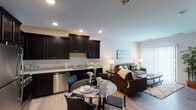 Located on S Adams St in Oswego, Illinois, this single-floor unit offers a thoughtfully designed open-concept layout that seamlessly connects the kitchen, dining, and living areas. The kitchen is a standout feature, showcasing rich dark cabinetry with raised-panel detailing, light granite countertops, and a full suite of stainless steel appliances including a gas range, microwave, refrigerator, and dishwasher. Light wood-style flooring runs throughout the main living area, enhancing the bright and contemporary ambiance. The dining area comfortably accommodates a round glass-top table, positioned between the kitchen and the spacious living room. The living room benefits from a large sliding glass door dressed with vertical blinds, allowing generous natural light to fill the space. The bedroom is a calm retreat featuring neutral carpet, a well-sized window with blinds, and ample room for furnishings. The bathroom carries the same dark cabinetry aesthetic with coordinating granite counters and wood-style flooring. Recessed lighting throughout and a neutral color palette give the unit a clean, modern feel overall.