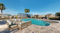 Located at 2700 Whitney Ave in Harvey, Louisiana, this outdoor amenity area offers a resort-style experience designed for relaxation and recreation. At its centerpiece is a generously sized freeform swimming pool with crystal-clear turquoise water, surrounded by an expansive travertine-tiled deck that provides ample space for lounging and socializing. Tall palm trees punctuate the landscape, lending a tropical ambiance beneath the open Louisiana sky.

The pool area is thoughtfully furnished with striped cushioned outdoor sofas and chairs, colorful lounge chairs, and shaded seating clusters with market umbrellas, creating comfortable gathering spots throughout the deck. A dedicated splash pad zone adjacent to the main pool features vibrant interactive water play structures, including tipping buckets and umbrella-style sprayers, making it an ideal space for younger visitors.

The perimeter is enclosed by a decorative fence that defines the space while maintaining an open, airy feel. A covered pavilion area with additional seating is visible in the background, offering a shaded retreat. The single-level layout ensures easy accessibility throughout. Overall, the space exudes a lively yet laid-back outdoor atmosphere well-suited for community enjoyment year-round.