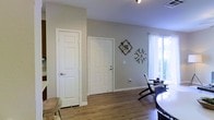 This single-floor residence presents a well-maintained interior with a cohesive and practical design. The main living area is outfitted with rich, dark hardwood-style flooring that extends throughout, providing a warm and grounded aesthetic. Crown molding lines the upper perimeter of the room, adding a subtle architectural detail that elevates the overall finish of the space. A ceiling fan with a built-in light fixture is centrally mounted, ensuring both comfort and illumination throughout the area. The entryway is defined by a solid white front door, framed neatly within the layout and flanked by clean white walls and trim. A hallway extends beyond the entry, suggesting additional rooms or utility spaces deeper within the floor plan. The neutral wall palette throughout creates a bright, open atmosphere that is versatile and easy to personalize. White baseboards and trim work complete the interior's cohesive look, tying together the flooring and walls with a crisp contrast. The overall ambiance of the space is tidy, understated, and move-in ready, making it a practical and comfortable single-floor living environment.