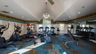 The Northland Passage Gym in Kansas City, MO offers a thoughtfully designed fitness space that combines functionality with an energizing atmosphere. The room features dramatic vaulted ceilings that rise to a striking peak, creating a sense of openness and volume throughout. Large windows and a prominent gabled window wall flood the interior with natural light while providing views of the surrounding greenery outdoors.

The fitness floor is covered in a vibrant patchwork-style carpet in teal, gold, and neutral tones, adding visual character to the space. An eye-catching motivational text border — featuring words like "Motivation," "Goals," "Achieve," "Commitment," and "Success" — runs along the upper walls in bold gold and black lettering, reinforcing an energizing workout environment.

The gym is comprehensively equipped with multiple treadmills, elliptical trainers, stationary and cycling bikes, and a cable/weight machine. A dedicated free weights area includes a full dumbbell rack and an adjustable bench, while a separate shelf unit holds medicine balls and stability balls for functional training. A ceiling fan and chandelier-style light fixture complement the architecture, and a water station is conveniently available. Large mirrors along one wall enhance the sense of space and allow users to monitor their form during workouts.