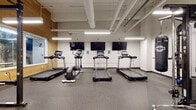 This fitness center is a comprehensively outfitted facility designed to accommodate a wide range of workout routines. The space is organized into distinct functional zones, each purpose-built to support different training modalities. The cardio area features multiple treadmills and an elliptical machine, each paired with a dedicated wall-mounted flat-screen television, allowing users to stay entertained during their workouts. Rubber flooring throughout the entire facility provides both durability and impact absorption.

The strength training zone is well-appointed with a multi-station cable machine, a power rack with an adjustable bench, and a full dumbbell rack spanning a broad weight range, complemented by a large wall mirror. Additional equipment includes a punching bag, stability balls, and a leg press machine, ensuring a thorough selection for resistance training.

Warm natural wood accent walls and large windows in the free weights area introduce an inviting aesthetic contrast to the otherwise industrial design. Exposed ductwork and overhead pipe systems run across the ceiling, reinforced by bright linear pendant lighting. A motivational typographic mural adorns one wall, and a water fountain station with a waste bin is conveniently accessible. The overall atmosphere is functional, clean, and energizing.