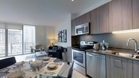 This single-floor space showcases a thoughtfully designed open-concept layout that seamlessly integrates the kitchen, dining, and living areas into one cohesive environment. The kitchen is outfitted with stainless steel appliances, including an electric range, over-the-range microwave, and dishwasher, complemented by rich, dark wood cabinetry that extends to the upper walls for ample storage. A neutral-toned countertop ties the culinary space together with a clean, modern aesthetic.

The dining area features a glass-top table with contemporary seating, positioned to take full advantage of the open sightlines across the space. The living area is furnished with a navy blue sofa and coordinating accent chairs arranged around a sleek coffee table, creating an inviting yet sophisticated lounge atmosphere. Framed abstract artwork adorns the walls, adding visual interest against the crisp white surfaces.

Floor-to-ceiling windows along the far wall flood the interior with natural light and provide views of the surrounding urban landscape, enhancing the airy, modern ambiance throughout the space.