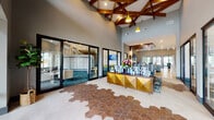 This single-story clubhouse is designed with a contemporary aesthetic that blends warm wood accents with bold, modern furnishings. The entry foyer makes an immediate impression with its striking hexagonal tile flooring in terracotta and cream tones, soaring ceilings with exposed wooden beam detailing, and pendant lighting in yellow and gold hues. A geometric gold reception desk anchors the space, while large glass sliding doors flood the interior with natural light and offer views to adjoining areas.

The lounge area features teal-patterned carpeting, textured stone-look wall cladding, and comfortable seating arranged around a fireplace with a mounted display. A café-style bar with a white marble countertop, bar stools, and dramatic pendant lighting serves as a casual gathering hub. A dedicated conference room offers a wood table surrounded by mustard-yellow chairs and a wall-mounted screen. A game room includes a pool table beneath sculptural wicker pendant lights. A cozy booth nook with slatted wood walls and tufted blue seating provides an intimate retreat. An illuminated mailbox center completes the facility's functional offerings.