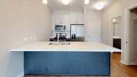This one-bedroom, one-bathroom unit is thoughtfully laid out across a single floor, making efficient use of its space. The main living area features warm hardwood-style flooring and neutral gray walls, complemented by track lighting that adds a modern, functional touch. Multiple electrical outlets are positioned along the baseboards, offering practical flexibility for furniture arrangement.

The kitchen is a standout feature, centered around a striking navy blue island with a white quartz countertop and an undermount sink. White upper cabinetry extends to the ceiling, providing ample storage, while a subway tile backsplash frames the stainless steel appliances, which include a gas range, built-in microwave, and a French door refrigerator with an ice and water dispenser.

The bedroom is carpeted for comfort and receives generous natural light through a large casement window with exterior views. The full bathroom includes a dark wood vanity with a white countertop and single sink, a wide mirror, and a glass-enclosed walk-in shower with white subway tile walls. Hardwood-style flooring continues into the bathroom, tying the space together cohesively.