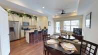 This single-floor, two-bedroom, one-bathroom residence offers a thoughtfully designed open-concept layout that maximizes its generous square footage. The kitchen is a standout feature, equipped with white cabinetry, stainless steel appliances including a range and over-the-range microwave, and light-toned granite countertops. A raised kitchen island with a rich, dark wood base provides bar-style seating for multiple guests, making it ideal for casual dining and entertaining.

The kitchen flows seamlessly into the dining area, which comfortably accommodates a full-sized table and chairs. Beyond the dining space, the living room is bright and welcoming, benefiting from a large double window and a glass-paneled door that brings in abundant natural light. A ceiling fan with a sleek black finish adds both style and functionality to the living area. Warm hardwood flooring runs throughout the main living spaces, creating a cohesive and inviting ambiance. Crown molding and recessed lighting add refined architectural details throughout, contributing to an overall atmosphere that is both comfortable and polished.