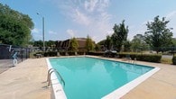 Located at 5201 Monaco Dr in Pascagoula, Mississippi, this single-story community space is defined by its generous outdoor amenities and relaxed Southern atmosphere. The centerpiece of the property is a large, rectangular outdoor swimming pool filled with clear, inviting turquoise water. The pool is equipped with metal entry railings for easy access and is surrounded by a wide concrete deck that provides ample space for lounging and recreation. Numerous pool-side chairs are arranged around the perimeter, offering comfortable seating for residents and guests. The pool area is enclosed by a chain-link fence, ensuring a secure and well-defined space. Mature trees line the surrounding landscape, providing natural shade and a lush, green backdrop that enhances the outdoor ambiance. The setting benefits from abundant natural light under open skies, creating a bright and welcoming environment. In the background, neighboring residential buildings are visible through the tree line, reflecting the community-oriented character of the property. Overall, the space presents a clean, functional, and inviting outdoor recreational environment well-suited to warm Gulf Coast summers.