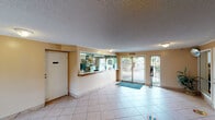 Located at 3360 W Lincoln Ave in Anaheim, California, this single-floor lobby serves as the primary reception and entry point for the property. The space is finished with light-colored square tile flooring throughout, lending a clean and practical aesthetic. Crown molding runs along the perimeter where the walls meet the textured ceiling, adding a subtle architectural detail to the otherwise straightforward interior.

The front desk area is prominently positioned and features a wide glass partition, allowing staff to maintain a clear line of sight across the lobby while remaining separated from the guest area. Behind the counter, organized shelving and administrative equipment are visible, indicating a fully operational reception setup.

Natural light enters through large glass doors and windows at the front entrance, illuminating the open floor plan and creating a bright, airy atmosphere. A blue entrance mat anchors the main doorway. An ATM machine and a brochure display rack are conveniently situated along one wall, providing guests with easy access to local information and financial services. A decorative potted plant near the entrance adds a touch of greenery to the space.

The lobby is open and uncluttered, offering ample room for guest circulation. Flush-mount ceiling fixtures provide supplemental lighting throughout, ensuring consistent illumination across the entire area.
