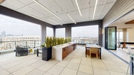 Located in Charlotte's vibrant NoDa arts district, this single-floor amenity space at 2226 North Davidson Street offers a thoughtfully designed retreat spanning a generous footprint. The layout comprises two distinct living areas, each with its own character and purpose.

The indoor lounge features striking slatted wood-panel accent walls and ceilings that lend a warm, contemporary aesthetic. Patterned geometric tile flooring adds visual interest underfoot, while recessed lighting and a wall-mounted television make the space both stylish and functional. A shuffleboard table and casual seating arrangements create an inviting social atmosphere, complemented by a wet bar with cabinetry.

The second living area transitions to a bright, open-concept space enclosed by expansive floor-to-ceiling windows on multiple sides, offering sweeping panoramic views of the Charlotte skyline and surrounding neighborhood. Linear LED lighting runs across a clean white ceiling, and tall live plants serve as natural dividers between a central kitchenette island with wood cabinetry and flexible dining and lounge seating.

A wraparound outdoor balcony with sleek glass-panel railings extends the space further, providing unobstructed views of the city and surrounding treetops. The covered portion of the balcony features a warm wood-plank ceiling with ceiling fans and a mounted television, ideal for year-round outdoor enjoyment. Large-format tile flooring flows consistently throughout, unifying all areas of the space.