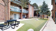 This inviting courtyard is nestled within a multi-story residential complex characterized by warm, multi-toned brick exteriors accented with vertical siding panels and teal-trimmed balconies. The buildings frame the courtyard on multiple sides, creating an enclosed, community-oriented atmosphere with a sense of privacy and openness simultaneously. The ground level features a generous stretch of well-kept artificial turf, lending the space a vibrant, green appearance year-round. Decorative natural boulders are placed throughout the lawn area, adding a landscaped, organic element to the design. Concrete walkways run alongside the buildings, providing clean pedestrian pathways through the courtyard. Black metal picnic tables are positioned throughout, offering comfortable outdoor gathering spots for residents. Mature pine and evergreen trees are scattered across the courtyard, providing shade and a natural canopy that softens the surrounding brick architecture. At one end of the complex, a gated outdoor swimming pool serves as a central amenity, enclosed by ornamental black iron fencing and accompanied by poolside seating and a red shade umbrella. Lush shrubs and potted plants enhance the pool area's landscaping. The overall ambiance is functional, communal, and well-maintained, offering residents a practical and pleasant outdoor environment.