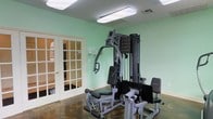 Located at 1733 Pops Ferry Rd in Biloxi, Mississippi, this single-floor fitness space offers a dedicated area for strength training and exercise. The room is finished with soft mint green walls that create a clean, energizing atmosphere, complemented by bright fluorescent overhead lighting that ensures excellent visibility throughout the space.

The centerpiece of the room is a multi-station cable and weight machine, providing a comprehensive strength training setup capable of targeting multiple muscle groups. The equipment includes a leg press attachment and adjustable bench, making it a versatile all-in-one workout station. The flooring features a durable dark surface well-suited for a fitness environment.

A set of classic French doors with glass pane inserts serves as the room's entry point, adding a refined architectural touch while allowing visibility into adjoining areas. The walls also feature utility controls and ventilation, contributing to the room's functional design. Overall, the space presents a practical and well-lit fitness environment suitable for a variety of strength training routines.
