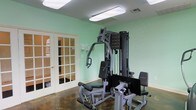 Located at 1733 Pops Ferry Rd in Biloxi, Mississippi, this single-floor fitness space offers a dedicated area for strength training and exercise. The room is finished with soft mint green walls that create a clean, energizing atmosphere, complemented by bright fluorescent overhead lighting that ensures excellent visibility throughout the space.

The centerpiece of the room is a multi-station cable and weight machine, providing a comprehensive strength training setup capable of targeting multiple muscle groups. The equipment includes a leg press attachment and adjustable bench, making it a versatile all-in-one workout station. The flooring features a durable dark surface well-suited for a fitness environment.

A set of classic French doors with glass pane inserts serves as the room's entry point, adding a refined architectural touch while allowing visibility into adjoining areas. The walls also feature utility controls and ventilation, contributing to the room's functional design. Overall, the space presents a practical and well-lit fitness environment suitable for a variety of strength training routines.
