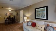 This single-floor space presents a comfortable and well-appointed living area that serves as the focal point of the home. The living room features warm hardwood flooring that extends throughout, contributing to a cohesive and inviting aesthetic. Neutral-toned walls provide a versatile backdrop, complemented by subtle wall-mounted décor including a large framed nature-inspired artwork that adds a vibrant visual element to the space.

The seating area is anchored by a generously sized light-colored sofa adorned with decorative pillows in rich jewel tones, adding warmth and personality. A classic table lamp on a side table provides soft ambient lighting, enhancing the room's cozy atmosphere. White-painted baseboards and door trim offer clean contrast against the warm wall tones.

A doorway visible from the living area leads further into the space, hinting at additional rooms beyond. A glimpse of a dining area with seating is visible in the background, suggesting an open and connected layout. The overall ambiance is warm, livable, and thoughtfully arranged, making it a practical and comfortable everyday living environment.