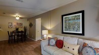 This single-floor space presents a comfortable and well-appointed living area that serves as the focal point of the home. The living room features warm hardwood flooring that extends throughout, contributing to a cohesive and inviting aesthetic. Neutral-toned walls provide a versatile backdrop, complemented by subtle wall-mounted décor including a large framed nature-inspired artwork that adds a vibrant visual element to the space.

The seating area is anchored by a generously sized light-colored sofa adorned with decorative pillows in rich jewel tones, adding warmth and personality. A classic table lamp on a side table provides soft ambient lighting, enhancing the room's cozy atmosphere. White-painted baseboards and door trim offer clean contrast against the warm wall tones.

A doorway visible from the living area leads further into the space, hinting at additional rooms beyond. A glimpse of a dining area with seating is visible in the background, suggesting an open and connected layout. The overall ambiance is warm, livable, and thoughtfully arranged, making it a practical and comfortable everyday living environment.