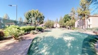This single-floor residence is situated within a thoughtfully landscaped community that offers a variety of outdoor amenities. The exterior environment is a standout feature, showcasing a well-maintained putting green surrounded by mature trees, manicured shrubs, and drought-tolerant plantings that thrive under the region's sunny skies. A tennis court enclosed by chain-link fencing is visible nearby, adding to the recreational appeal of the surroundings.

The building exterior presents a stucco finish in a neutral tone, consistent with the Mediterranean-inspired architecture common to the community. A concrete staircase with metal railings leads to upper-level units, while ground-floor entry points are accessible directly from the shared walkways. The entryway to the unit is framed by a gated passage, with climbing vines adding a touch of greenery to the approach.

Interior details visible suggest smooth, light-colored walls throughout, contributing to a clean and neutral aesthetic. The unit's single-floor layout offers straightforward accessibility. The surrounding community infrastructure, including well-kept pathways and lush tree canopy, creates a calm and pleasant outdoor ambiance that complements the living experience within this residential setting.