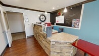 Located at 1195 Western Ave in Beaver, PA, this single-floor community space offers a thoughtfully designed interior that blends comfort and functionality. The space is anchored by a warm lounge area featuring rich hardwood flooring, decorative columns with dark crown molding, and a striking stone-clad fireplace wall fitted with built-in shelving and a mounted television. Comfortable seating, including upholstered sofas and accent chairs in neutral and bold red tones, creates an inviting atmosphere for gathering and relaxation. Natural light filters in through multiple windows dressed with dark curtain panels, enhancing the bright, neutral-toned walls. A stone-faced service counter with a granite top serves as a central hub, seamlessly connecting the lounge to a casual café dining area. The café section features vibrant teal accent walls, a chalkboard menu display, pendant lighting, and an assortment of dining tables and chairs arranged to accommodate multiple guests. The open layout allows the two zones to flow naturally into one another, making the space versatile and community-oriented. Recessed lighting throughout ensures consistent illumination across the entire floor.