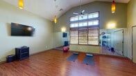 Located on Riley Road in Easton, PA, this single-floor fitness and yoga studio is purpose-built for movement and wellness. The space is defined by its striking vaulted ceilings, which rise dramatically and lend an open, airy quality throughout. Warm hardwood flooring spans the entire room, providing a durable and attractive surface well-suited for yoga, stretching, and general exercise.

Large windows with wood-trimmed blinds line one wall, filling the studio with generous natural light, while a bank of transom windows above further amplifies the brightness and enhances the sense of height. A ceiling fan centered beneath the peak of the vault ensures comfortable air circulation during active use.

Full-length wall mirrors run along two walls, a practical and professional feature that allows users to monitor form and technique. The walls are painted in a soft sage green, contributing to a calm, focused atmosphere. Pendant lights with amber glass shades add a warm, inviting glow to complement the natural light.

The studio is equipped with yoga mats, stability balls, resistance accessories, and a storage rack for fitness equipment. A wall-mounted flat-screen television and an audio unit provide multimedia support for classes or individual sessions. A secondary door offers additional access, making the space functional for both instructors and participants.