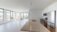 This thoughtfully designed single-floor residence showcases a clean, contemporary aesthetic throughout. The open-concept kitchen and living area forms the heart of the unit, anchored by a generous granite-topped island with an integrated sink and a sleek pendant light overhead. The kitchen is equipped with stainless steel appliances, including a range, microwave, and refrigerator, complemented by tall white cabinetry and dark countertops along the back wall. Light wood-tone flooring flows seamlessly throughout the entire unit, enhancing the bright, airy atmosphere.

The living area is flooded with natural light through floor-to-ceiling windows and sliding glass doors that open to a balcony, offering expansive views of the surrounding cityscape and treetops. Additional bedrooms feature large windows that frame neighborhood and skyline views, with ample sunlight and a wall-mounted mini-split system providing climate control.

Both bathrooms maintain the unit's consistent design language, featuring granite-topped white vanities, large mirrors, and wood-grain doors. One bathroom includes a walk-in shower with white tile surround, while the second offers a long double-sink vanity with generous storage. The overall ambiance is modern, minimalist, and light-filled throughout.