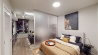 This contemporary single-floor residence showcases a smart, open-concept layout that maximizes every square foot with intentional design. The kitchen is a standout feature, outfitted with sleek grey cabinetry, stainless steel appliances including a full refrigerator, range, microwave, and dishwasher, complemented by a white subway tile backsplash, light stone countertops, and a matte black sink fixture. Open shelving adds both style and functionality, while a large window frames city views, bringing in additional light and ambiance. The living area flows seamlessly from the kitchen, offering a versatile space that accommodates both a comfortable seating arrangement and a dedicated workspace or study nook. A freestanding open shelving unit serves as a stylish room divider, adding visual interest and organizational utility. The sleeping area is cleverly separated from the living space by a full-width frosted sliding glass partition, providing privacy without sacrificing the open feel of the floor plan. The bedroom features a built-in closet with organized shelving. Light wood-style flooring runs throughout, tying the spaces together cohesively. The neutral color palette, modern fixtures, and clean architectural lines create a polished, urban ambiance throughout.