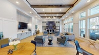 This single-floor amenity center is thoughtfully designed to blend style and functionality. The centerpiece is a generous clubroom featuring soaring coffered ceilings adorned with rich dark wood beams, a striking globe chandelier, and warm hardwood flooring. Multiple seating arrangements—including patterned accent chairs, a plush sofa, and bistro-style tables—create versatile spaces for socializing or relaxing. A wall-mounted TV and built-in cabinetry add convenience, while floor-to-ceiling windows flood the room with natural light and offer views of an outdoor pool and patio area.

The kitchenette area is a visual standout, showcasing a bold navy herringbone tile accent wall, soft gray shaker cabinetry, open wood shelving, white countertops, and a stainless steel refrigerator—ideal for casual gatherings.

The expansive fitness center is outfitted for comprehensive workouts, featuring strength training machines, a power rack, free weights, kettlebells, medicine balls, cardio equipment including treadmills, stair climbers, and stationary bikes, plus large wall mirrors and a mounted flat-screen TV. Consistent with the clubroom, dark wood beams and tray ceilings with ceiling fans carry the design theme throughout. Additional areas include a neatly appointed hallway with bistro seating and restroom access, completing this well-rounded amenity facility.