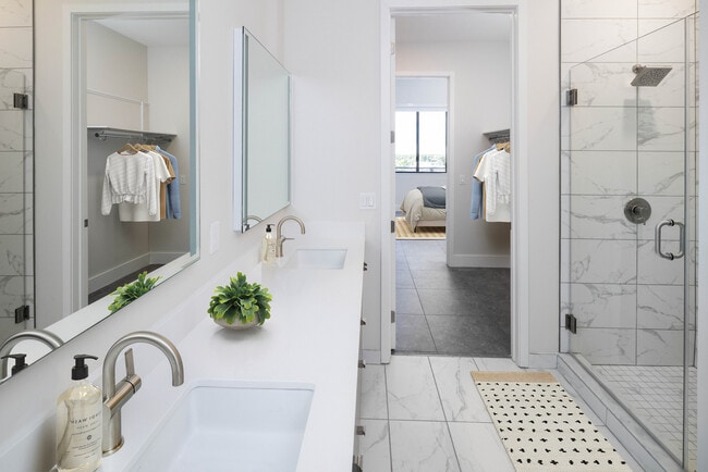 Baño Signature Collection con gabinetes grises, encimera de cuarzo blanco, espejos de tocador iluminados, mampara de ducha de porcelana y piso de baldosas de porcelana. - Avalon South Miami