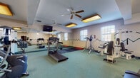 Located at 11250 Somerset Dr in North Royalton, Ohio, this fitness center is a comprehensively equipped single-floor exercise facility designed to support a full range of workout routines. The space is organized into distinct functional zones, accommodating both cardiovascular training and strength conditioning.

The cardio area features multiple treadmills, elliptical trainers, upright stationary bikes, and stair-stepping machines, providing users with a variety of aerobic exercise options. The strength training section includes a Vectra multi-station cable machine, a Cybex resistance unit, and additional weight benches and attachment stations suited for targeted muscle conditioning.

The flooring throughout the space consists of durable green rubber gym flooring, with rubber mat sections placed beneath equipment for additional protection and traction. Full-length wall mirrors span one side of the room, enhancing the sense of openness and allowing users to monitor their form during exercise. A wall-mounted flat-screen television is positioned for visibility across the main workout area.

Overhead, coffered ceiling details add architectural interest, while multiple ceiling fans and warm rectangular light fixtures provide consistent air circulation and illumination. Natural light enters through several windows along the perimeter walls. Decorative zigzag accent lines run along the walls, adding a subtle visual element to the overall design.