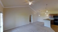 This single-floor residence showcases an open and airy design defined by impressive vaulted ceilings that lend a sense of grandeur to the main living area. A ceiling fan with a light fixture is centrally positioned, ensuring both comfort and illumination throughout the space. Crown molding adds a refined architectural touch along the upper walls, complementing the clean, neutral color palette that makes the interior feel bright and welcoming.

The living area features plush wall-to-wall carpeting that adds warmth underfoot and flows seamlessly throughout the space. A doorway connects the main living area to an adjoining room, providing a natural sense of transition and privacy. To one side, a partial view of the kitchen is visible, where a pendant light hangs above a countertop overhang, hinting at a dining or bar-style seating arrangement that promotes casual interaction between the kitchen and living spaces.

The soft, neutral tones on the walls and ceiling, combined with the open layout and elevated ceiling height, create a calm and versatile environment well-suited for both relaxed living and entertaining. The overall ambiance is clean, spacious, and move-in ready, offering a comfortable and functional single-level living experience.