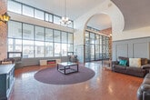 Located at 3020 Glencrest Road in Burlington, Ontario, this distinctive living space makes a bold architectural statement from the moment you enter. The room is defined by its impressive double-height ceilings, which lend a grand, open sense of volume rarely found in a single-floor layout. Warm terracotta tiles laid in a classic herringbone pattern span the entire floor, anchoring the space with rich, earthy tones.

A full-height curtain wall of steel-framed windows dominates one side of the room, flooding the interior with abundant natural light and offering clear views of the surrounding neighbourhood streetscape. Anchoring the window wall is a sleek, modern electric fireplace unit, providing a striking contrast of contemporary comfort against the room's more traditional elements.

Exposed brick walls add texture and warmth to the space, accented by vintage-style wall lanterns that evoke a classic charm. An ornate wrought-iron lattice partition with a decorative arch serves as an elegant interior divider, while a curved mezzanine balcony overhead features intricate ironwork railings, reinforcing the space's sophisticated character.

The living area is furnished with leather sofas and paired lounge chairs, arranged around a central circular area rug. A modular open-shelf unit and a vibrant blue sideboard complete the functional yet characterful design of this remarkable communal space.