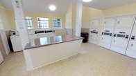 Located at 9475 W Sam Houston Pky S in Houston, Texas, this single-story community laundry facility offers a clean, functional environment designed for convenience. The space features vaulted ceilings that lend an open, airy feel, complemented by soft yellow walls and neutral tile flooring throughout. Natural light filters in through multiple windows along one wall, brightening the interior and creating a welcoming atmosphere. A central granite-topped folding counter serves as a practical workspace, providing ample surface area for sorting and folding laundry. Along the perimeter, stacked washer and dryer units are neatly arranged and equipped with coin-operated controls, offering multiple machines to accommodate residents efficiently. A structural column adds an architectural element to the space without disrupting the overall flow. Additional features include a utility area with washing machines along the windowed wall, a waste bin for easy disposal, and a door providing access to adjacent areas. The space is well-lit with ceiling-mounted light fixtures and maintains a tidy, organized layout. Overall, this laundry facility is thoughtfully designed to be both practical and comfortable for everyday residential use.