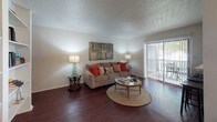 This single-floor residence is thoughtfully laid out to maximize comfort and functionality across its key living spaces. The living room serves as the heart of the home, featuring rich dark hardwood flooring, a textured ceiling, and a large sliding glass door fitted with vertical blinds that opens onto a private outdoor patio — allowing an abundance of natural light and pleasant outdoor views to fill the space. The kitchen is equipped with warm wood cabinetry, a double stainless steel sink, ample counter space, and a window overlooking lush greenery, making it both practical and inviting. Adjacent to the kitchen, the dining area accommodates a round table with seating for four, accented by a ceiling fan overhead and connected seamlessly to the main hallway. The bedroom is a serene retreat, carpeted for warmth and comfort, with natural light filtering through a large window dressed with elegant curtains and blinds. A closet and nightstand with soft lamp lighting complete the space. The bathroom features a wood-toned vanity with a large mirror, overhead lighting, and a tub-shower combination with a decorative curtain. Throughout the residence, neutral wall tones and cohesive finishes create a warm, welcoming ambiance.