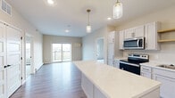 This well-appointed single-floor residence offers 2 bedrooms, 2 bathrooms, and 3 closets within a thoughtfully designed layout. The open-concept kitchen and living area serves as the heart of the home, featuring light hardwood-style flooring, recessed lighting, and two stylish brass pendant lights that anchor the central island with a clean white quartz countertop. The kitchen is fully equipped with white shaker cabinetry, stainless steel appliances including a refrigerator, electric range, microwave, and dishwasher, as well as an open wood shelf for additional display storage. The living area flows naturally toward sliding glass doors that open onto a private balcony, bringing in generous natural light and outdoor views of the surrounding neighborhood. The bedrooms are carpeted for comfort, each with access to natural light through windows offering exterior views. The primary bedroom connects conveniently to its own bathroom area with dedicated closet space. Both bathrooms are finished with white shaker-style vanity cabinetry, light countertops, hardwood-style flooring, and contemporary black hardware accents. Baseboard heating is present throughout, and a wall-mounted HVAC unit serves the living area. The space maintains a clean, neutral color palette throughout, creating a bright and cohesive aesthetic.