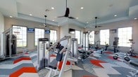 This single-story fitness center is designed to offer a comprehensive workout environment with a clean, modern aesthetic. The space is anchored by a tray ceiling with recessed lighting and multiple ceiling fans that provide both visual depth and effective air circulation throughout the room. Large multi-pane windows line the walls on multiple sides, flooding the interior with natural light and creating an open, airy atmosphere.

The flooring is a standout design element, featuring a bold geometric pattern of interlocking shapes in gray, white, and red-orange, adding energy and visual interest to the space. Strength training equipment, including cable machines and free weight stations, is centrally positioned, while cardio equipment such as stationary bikes and benches occupies the perimeter areas. Wall-mounted mirrors along one side enhance the sense of depth and allow users to monitor their form during workouts.

Multiple flat-screen televisions are mounted at various points around the room, ensuring visibility from most areas of the floor. The light gray walls complement the contemporary equipment and flooring palette, creating a cohesive, motivating environment well-suited for a variety of fitness activities.