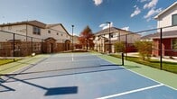 Located at 1841 Mansfield Webb Rd in Arlington, Texas, this outdoor recreational court is a well-appointed community amenity designed for active play and social engagement. The court features a regulation-sized layout with a crisp two-tone surface — a blue playing field accented by green out-of-bounds zones — with clearly marked white boundary lines and a center net, making it suitable for both pickleball and tennis.

The court is fully enclosed by a sturdy chain-link fence that ensures safety and ball retention during play. Tall light poles with solar-assisted or electric fixtures are positioned around the perimeter, allowing for evening use and extending playtime well beyond daylight hours. A dedicated entry gate provides easy access to the court.

Surrounding the court is a neatly landscaped green area featuring maintained grass, young trees, and mulched garden beds. A striking red pergola structure is visible just beyond the court's boundary, offering a shaded gathering spot for spectators or players resting between matches. The area is bordered by a warm cedar privacy fence, with a backdrop of two-story residential homes that give the space a community-oriented character. Open skies above provide an abundance of natural light, creating an inviting and energetic atmosphere for outdoor recreation.