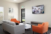 Located at 51 E Blue Heron Ln in Meridian, Idaho, this single-story community clubhouse offers a well-rounded gathering space designed for both relaxation and activity. The entry foyer sets a welcoming tone with wood-look flooring, a tray ceiling with warm gold accents, and ample natural light filtering through flanking windows alongside the glass-paneled front door.

The main lounge area is generously proportioned, featuring patterned carpet tiles in neutral gray tones that run throughout much of the space. Bold orange accent chairs and a white sofa are arranged around a central coffee table, creating an inviting seating arrangement. A wall-mounted flat-screen TV and exterior door access to an outdoor patio further enhance the room's functionality. Colorful abstract artwork adds a creative touch to the space.

The kitchen is fully equipped with dark walnut-toned cabinetry, black appliances including a range, microwave, and refrigerator, and light-toned countertops. A peninsula with bar seating provides a casual dining or meeting area.

A dedicated fitness room includes cardio equipment such as treadmills, ellipticals, and a power tower, lit by natural light from multiple windows. Dual drinking fountains are conveniently located nearby. The overall design is clean, modern, and functional, making this clubhouse a practical community amenity.
